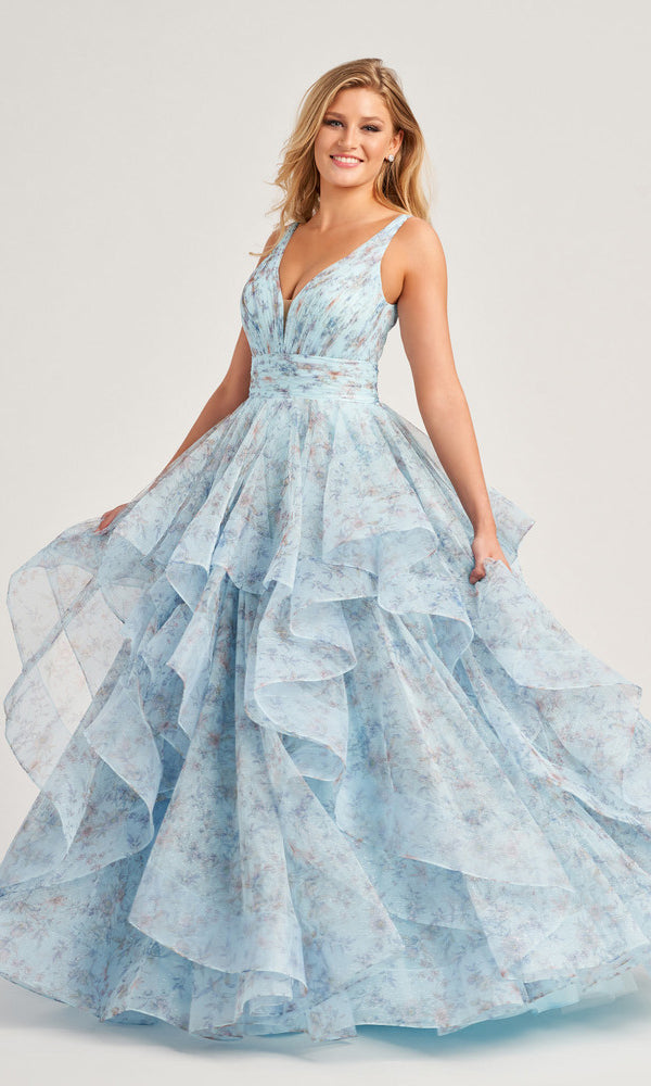 A woman smiles and stands against a plain background wearing the Colette Tiered Long Print Prom Ball Gown CL5273—a light blue, floral print dress featuring a sleeveless deep V-neck and a long tiered ball gown skirt.